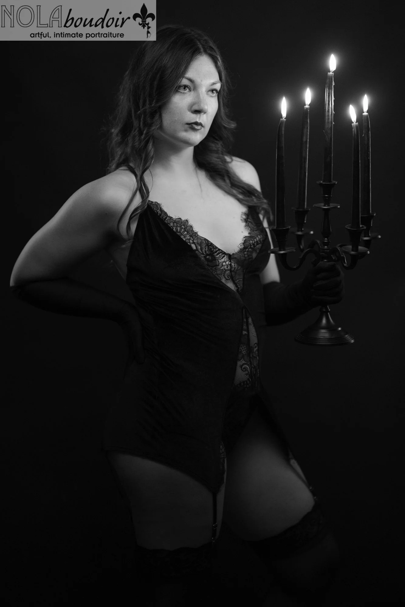A woman in black lingerie and gloves poses with a lit candelabra against a dark backdrop under moody, dramatic studio lighting.