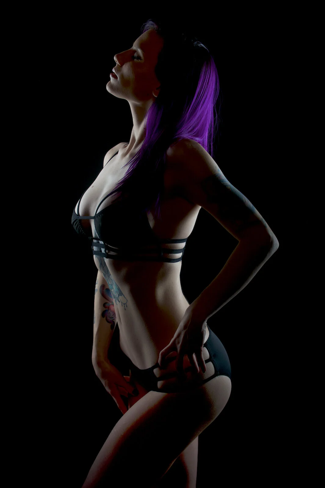 A purple-haired woman in strappy black lingerie poses in profile under dramatic rim lighting against a black background.