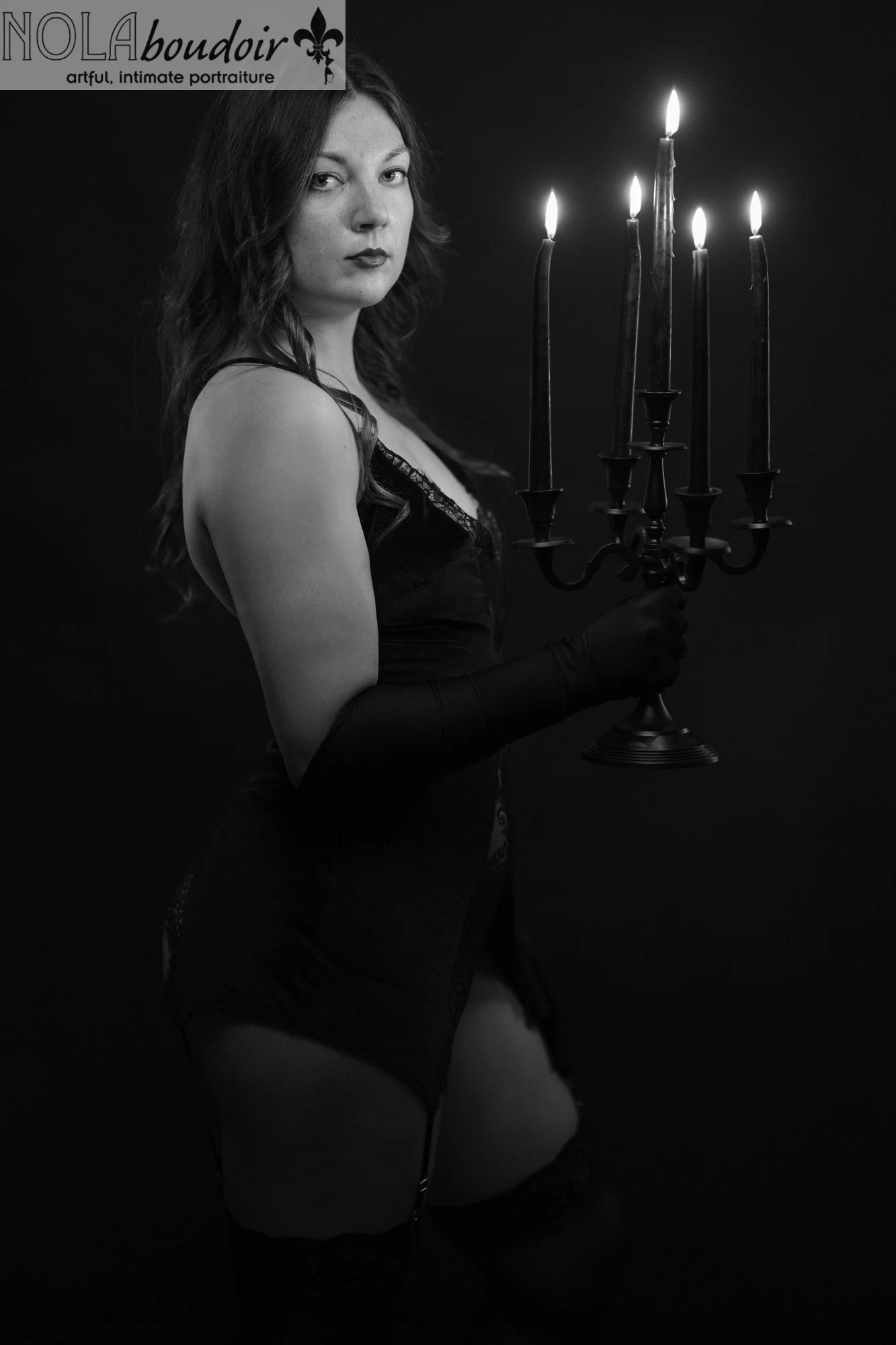 Woman in black lingerie and satin gloves holds a lit candelabra against a dark backdrop in moody, dramatic lighting.