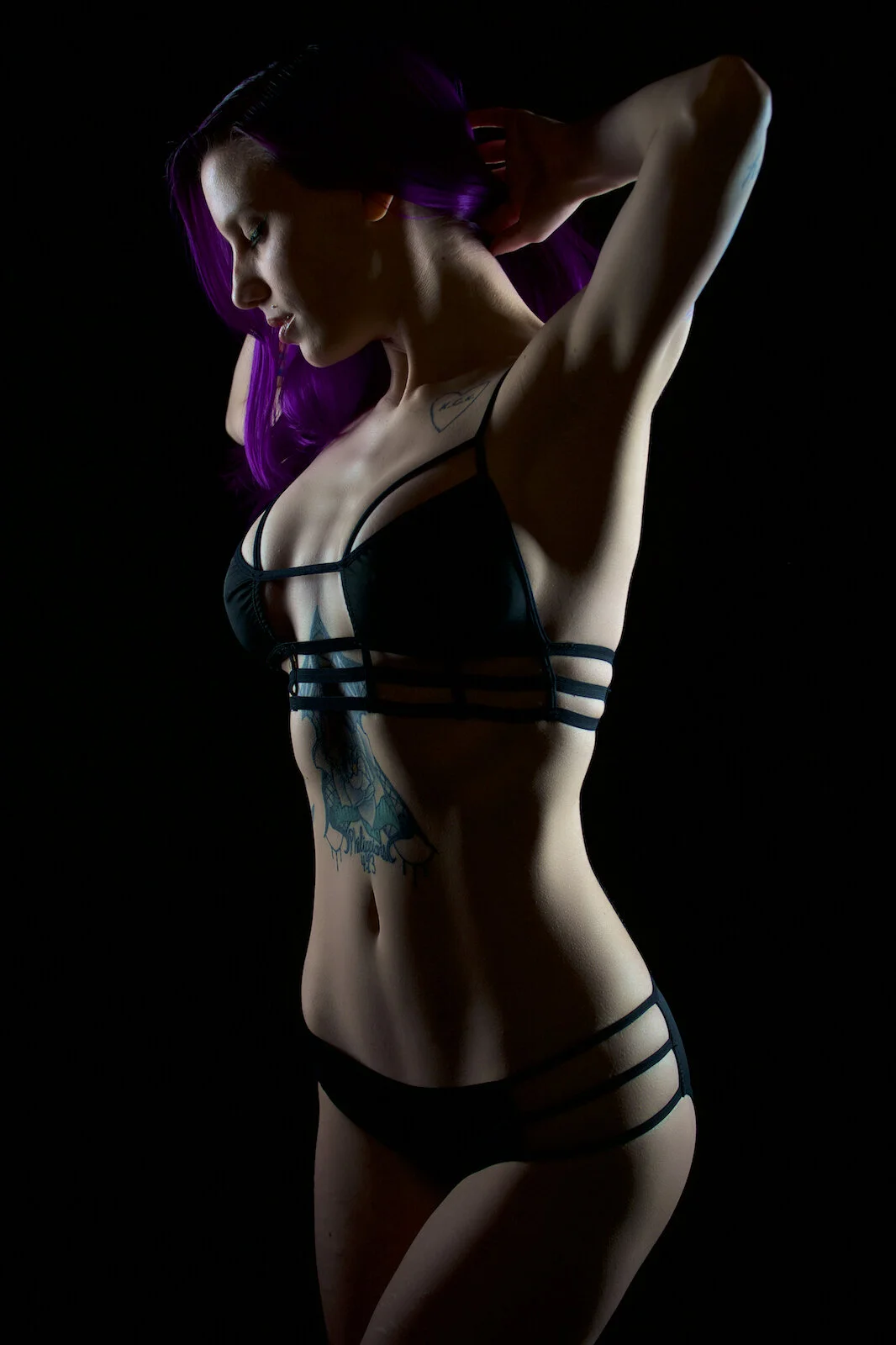 Woman with purple hair poses with arms raised in strappy black lingerie against a dark backdrop with moody rim lighting.
