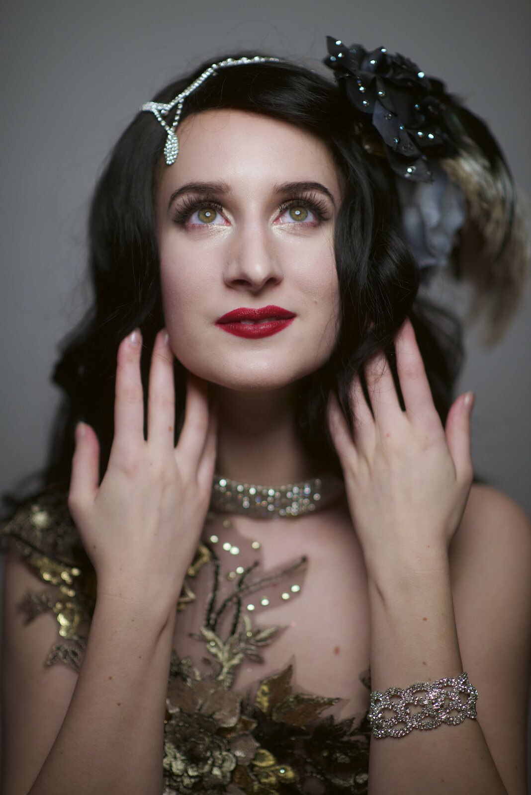Woman in vintage gold lace and crystal headpiece gazes upward dreamily with hands framing her neck under soft light.